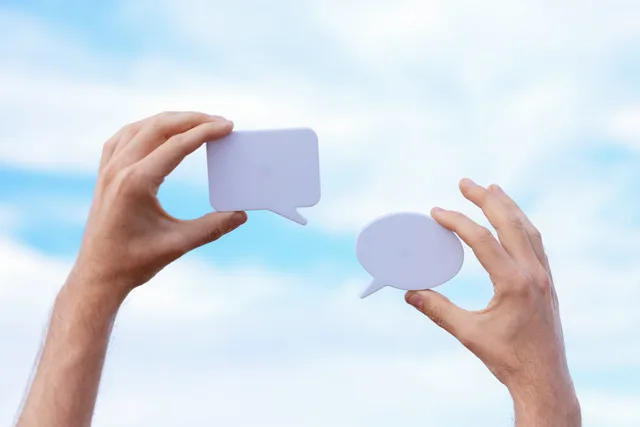 Close-up of hands holding speech bubbles, symbolizing communication and dialogue against a blue sky. Scrivere contenuti social chiari
